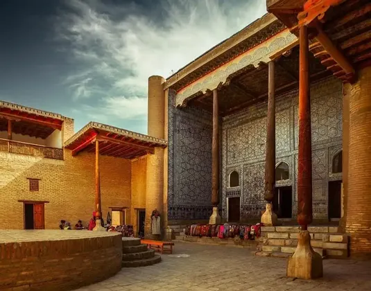 Un cortile soleggiato con edifici in mattoni, caratterizzato da grandi pilastri in legno intagliato e una facciata decorata con intricate piastrelle geometriche blu.