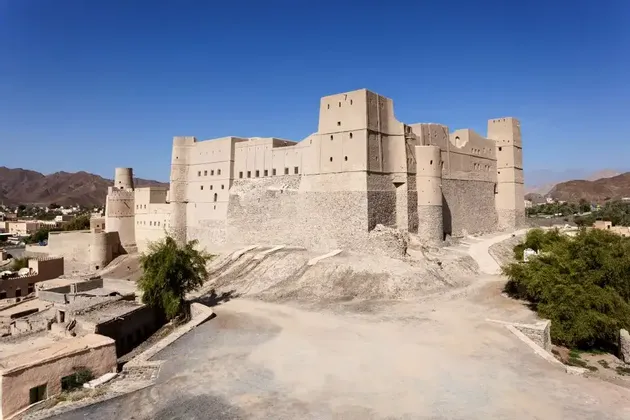 Una grande fortezza color sabbia con torri sorge su una collina che domina una città e montagne sotto un limpido cielo azzurro.
