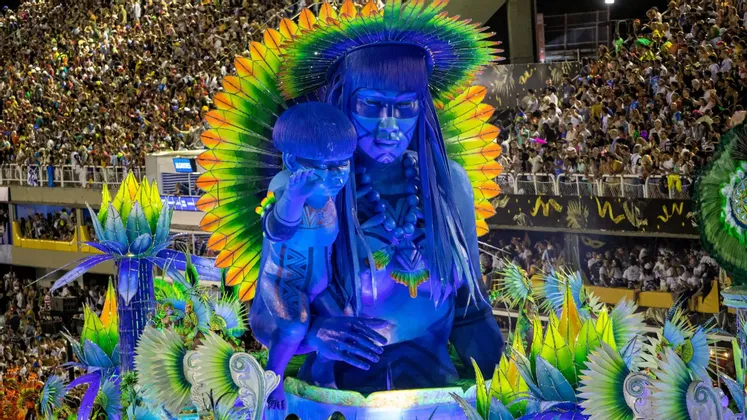 Ein großer, blauer Karnevalswagen mit einer indigenen Frau und einem Kind mit farbenprächtigem Federkopfschmuck, bei einem Umzug vor einer großen Menschenmenge.