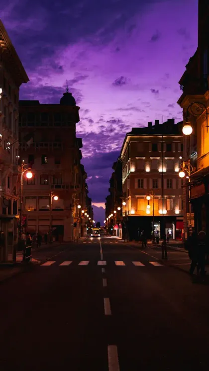Una strada cittadina fiancheggiata da edifici illuminati e lampioni sotto un vibrante cielo crepuscolare viola.