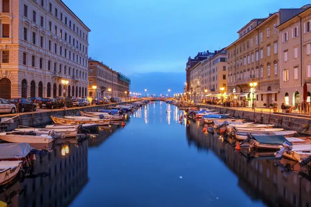 Piccole imbarcazioni sono ormeggiate lungo un canale cittadino al crepuscolo, con edifici illuminati che si riflettono nell'acqua calma.
