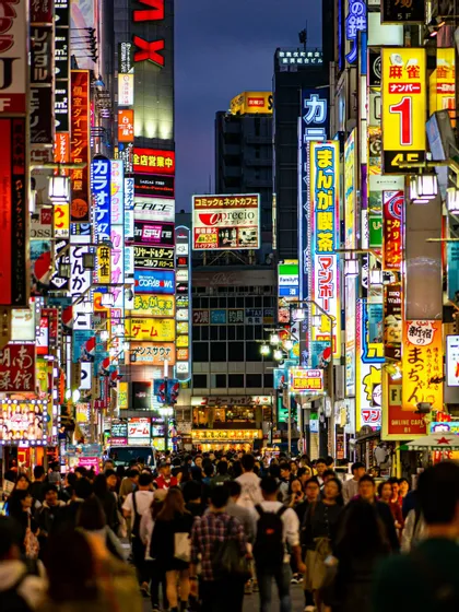 Una folla di persone cammina lungo una stretta strada cittadina al crepuscolo, illuminata dal bagliore di numerose e colorate insegne al neon giapponesi sugli edifici.