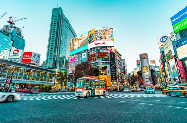 Un autobus colorato su un attraversamento pedonale in un trafficato incrocio cittadino, circondato da grattacieli e grandi cartelloni digitali con caratteri giapponesi.