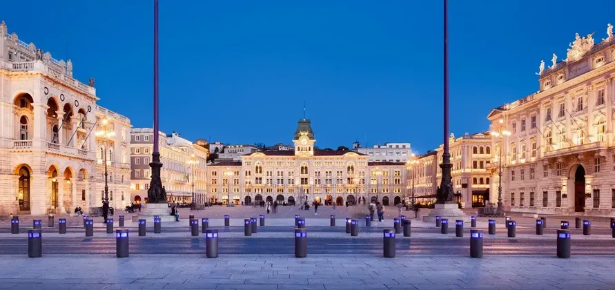Una vasta piazza cittadina, circondata da edifici storici illuminati, sotto un cielo blu intenso al crepuscolo.