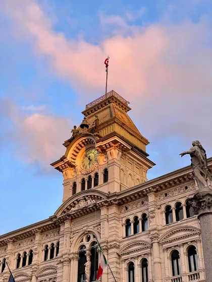 Uno scatto dal basso di un'ornata torre dell'orologio su un edificio storico, contro un cielo con nuvole rosa al tramonto.