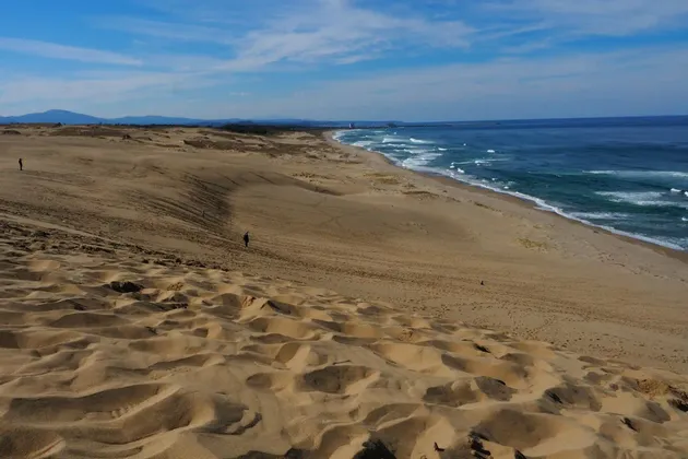 Vaste dune di sabbia che conducono a una costa oceanica blu, con alcune figure distanti che camminano sotto un cielo parzialmente nuvoloso.