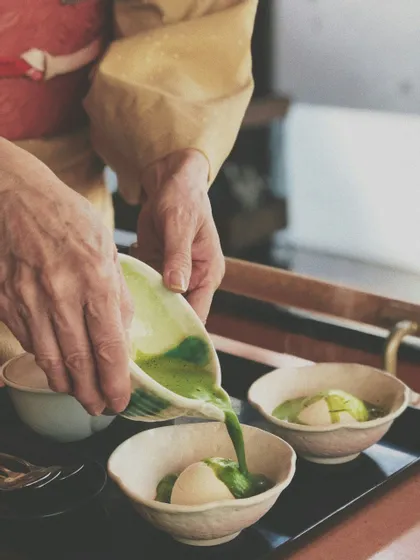Una persona in kimono giallo versa salsa matcha verde da una ciotola di ceramica su palline di gelato alla vaniglia.