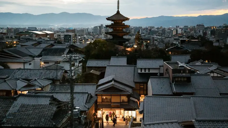 Una pagoda alta svetta su un panorama cittadino di tetti tradizionali al crepuscolo, con montagne lontane sotto un cielo nuvoloso.