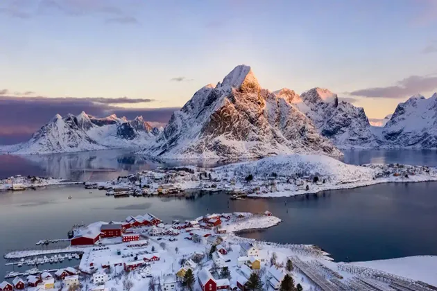 Eine Luftaufnahme eines verschneiten Küstendorfes mit roten Häusern, gelegen am Fuße großer, schneebedeckter Berge bei Sonnenaufgang.