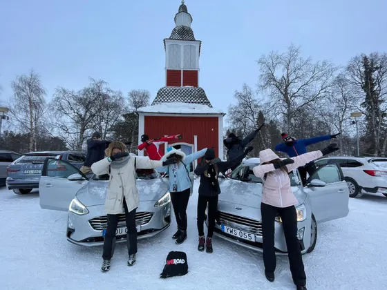Eine WeRoad-Gruppe posiert im 'Dab'-Stil neben zwei Autos in einer verschneiten Landschaft, mit einem roten Gebäude im Hintergrund.