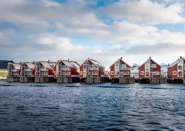Eine Reihe roter Stelzenhäuser steht über dunkelblauem Wasser, ihre Dächer sind leicht mit Schnee bedeckt.
