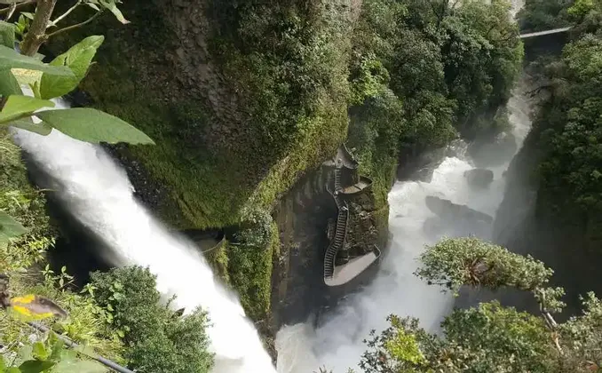 Una vista dall'alto di una grande cascata che si getta in una gola verde lussureggiante, con una scala in pietra tortuosa sulla parete rocciosa.