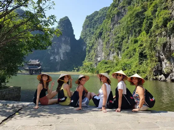 Un viaje en grupo de WeRoad con seis mujeres con sombreros cónicos sentadas en un muelle de piedra junto a un lago, con una pagoda y verdes montañas kársticas de fondo.