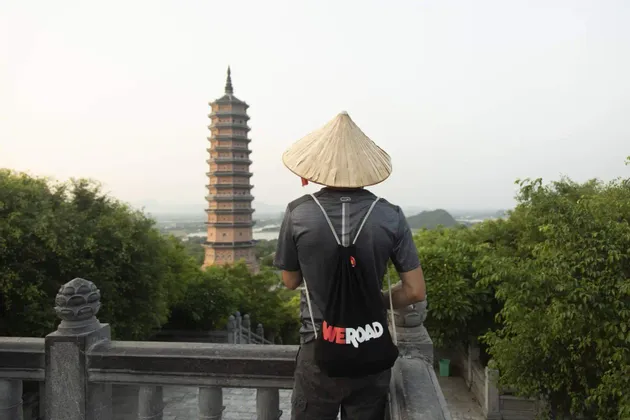 Un hombre de espaldas, con sombrero cónico y una mochila WeRoad, de pie en un balcón de piedra con vistas a una alta pagoda.