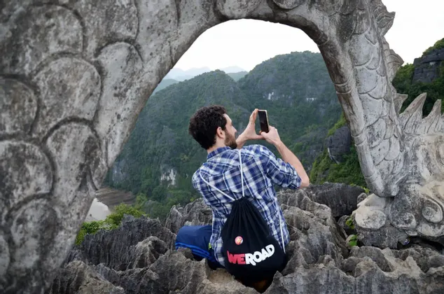 Un hombre con una mochila WeRoad se sienta en un mirador rocoso, tomando una foto de las montañas a través de un arco de dragón de piedra.
