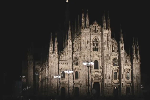 The intricate facade of a large Gothic cathedral with many spires, illuminated by lights against a dark night sky.
