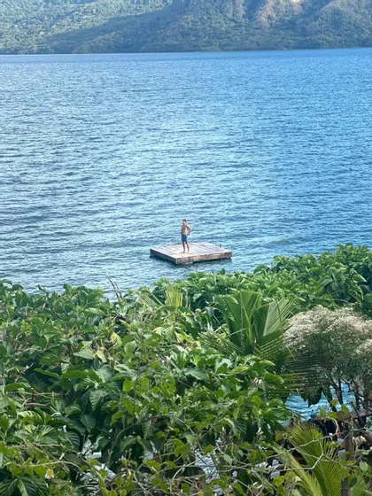 Un homme en short se tient sur un radeau en bois flottant sur un grand lac, entouré de montagnes vertes et d'une végétation luxuriante.