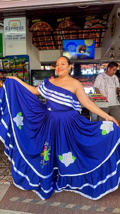 Une femme souriante pose, déployant les côtés de sa robe bleue traditionnelle à une épaule, agrémentée de rubans blancs et de broderies florales.