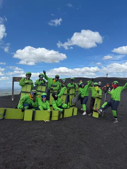 Un voyage de groupe WeRoad en combinaisons vert vif et lunettes posant avec des planches sur une sombre pente volcanique sous un ciel bleu.