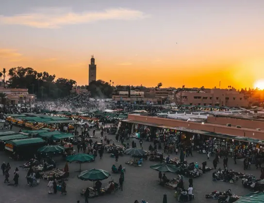 Una vista dall'alto di una vivace piazza cittadina piena di bancarelle e persone al tramonto, con un'imponente torre in lontananza.