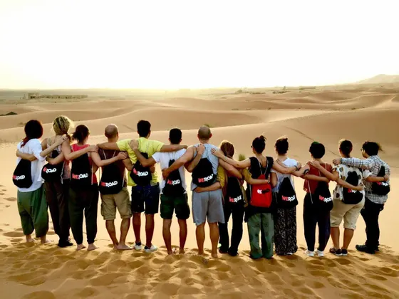 Eine WeRoad-Gruppe steht Arm in Arm auf einer Sanddüne und blickt bei Sonnenuntergang über eine weite Wüste.