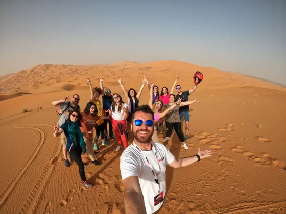 Ein Mann macht ein Selfie mit einer WeRoad-Reisegruppe, die auf großen Sanddünen in einer Wüste posiert und lächelt.