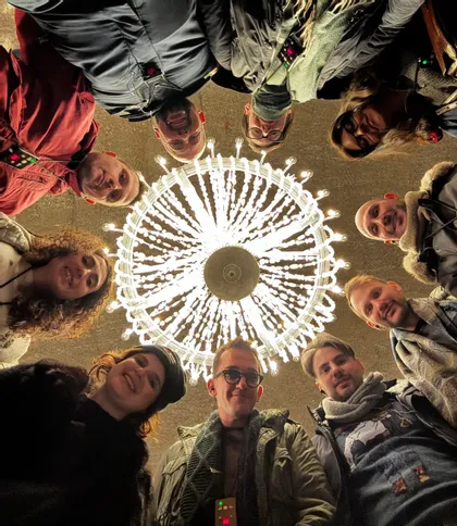 Un gruppo WeRoad con persone sorridenti in cerchio, visto dal basso, con un grande lampadario illuminato al centro.