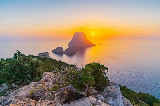 Un atardecer dorado sobre el mar y un gran islote rocoso, visto desde un acantilado verde con una torre de piedra.