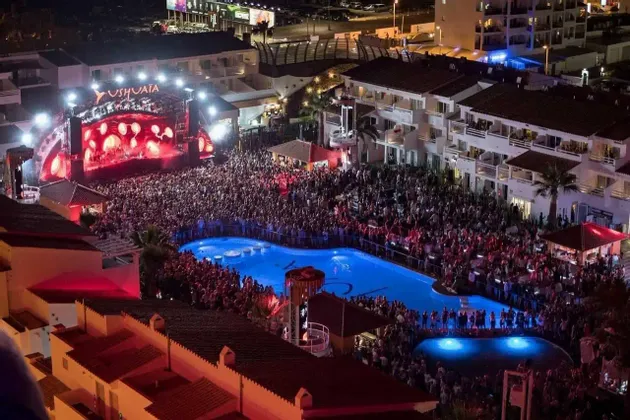 Una vista aérea de una enorme multitud en un concierto nocturno al aire libre, apiñada alrededor de un escenario muy iluminado y una piscina azul brillante.