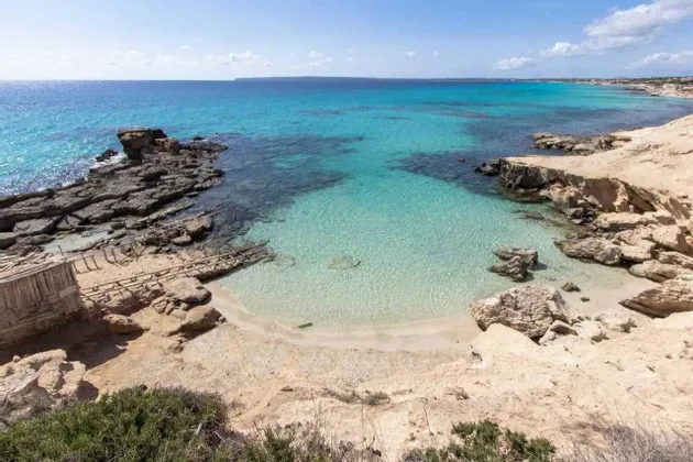 Una vista aérea de una cala apartada con agua turquesa cristalina, una pequeña playa de arena y acantilados rocosos bajo un cielo azul.