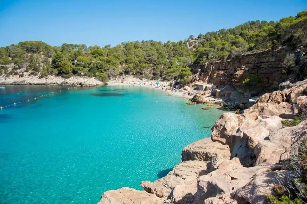 Una vista desde un acantilado rocoso de una cala apartada con agua turquesa cristalina, una playa de arena con gente y una costa arbolada.