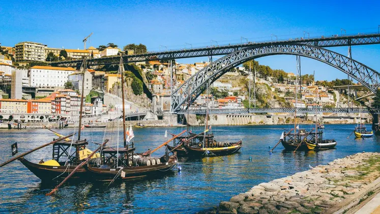 Barche tradizionali in legno galleggiano su un fiume con un grande ponte ad arco in metallo e una città collinare sullo sfondo.