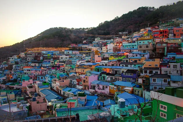 A dense hillside village of colorful houses with bright blue, pink, and green roofs, viewed at sunset.