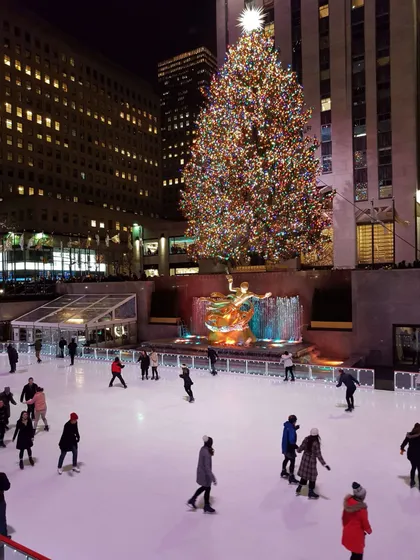 Des gens patinant sur glace la nuit sur une patinoire extérieure devant un grand sapin de Noël illuminé et des gratte-ciel de la ville.