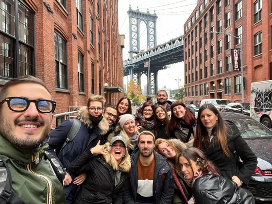 Un groupe WeRoad en voyage sourit pour un selfie dans une rue urbaine, entre des bâtiments de briques, avec un grand pont en acier en arrière-plan.