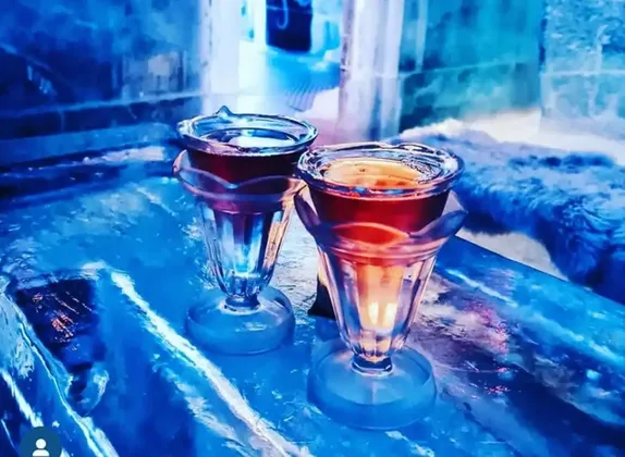 Dos vasos de chupito hechos de hielo, llenos de una bebida roja, reposan sobre una barra de bar también hecha de hielo con iluminación azul.