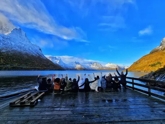 Un grupo de viaje de WeRoad está en un muelle de madera con las manos levantadas, contemplando montañas nevadas al otro lado de un lago bajo un cielo azul.