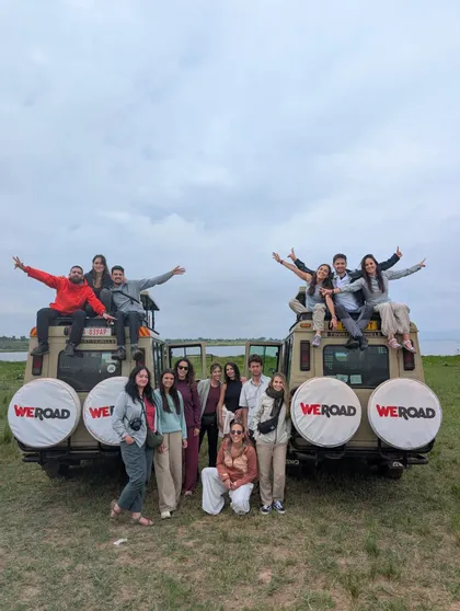 Un gruppo WeRoad in viaggio posa per una foto di fronte e sopra due jeep da safari in un campo erboso sotto un cielo coperto.