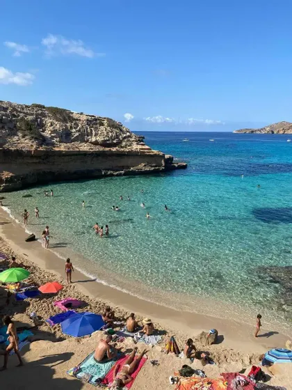 Gente tomando el sol en una playa de arena y nadando en las claras aguas turquesas de una cala rocosa en un día soleado.