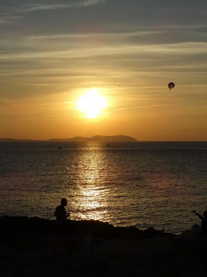 Siluetas de personas en una costa rocosa observan un atardecer dorado sobre el mar, con un parapente visible en el cielo.