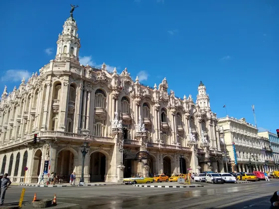 Un edificio classico, ornato e di colore chiaro, con torri, si erge all'angolo di una strada cittadina. Davanti, sotto un cielo azzurro, sono parcheggiati taxi gialli d'epoca e altre auto.