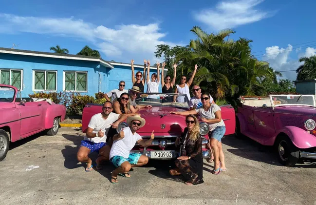 Un gruppo WeRoad in viaggio sorride e posa per una foto intorno a una decappottabile rosa d'epoca, davanti a un edificio blu.