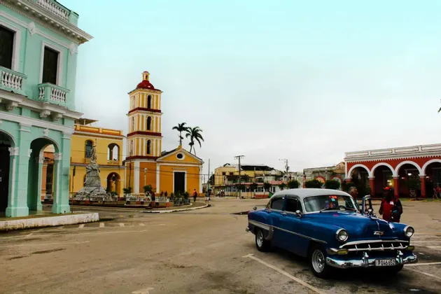 Un'auto d'epoca blu parcheggiata in una piazza, davanti a una chiesa gialla e ad edifici coloniali colorati.