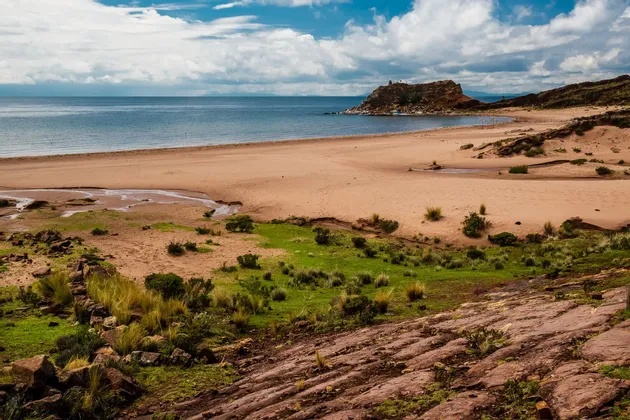 Una playa amplia y arenosa junto a un cuerpo de agua tranquilo, con zonas de césped verde y un promontorio rocoso bajo un cielo parcialmente nublado.