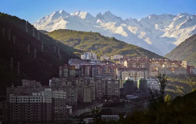 A city of apartment buildings built into a green mountain valley with a large, snow-capped mountain range in the background.