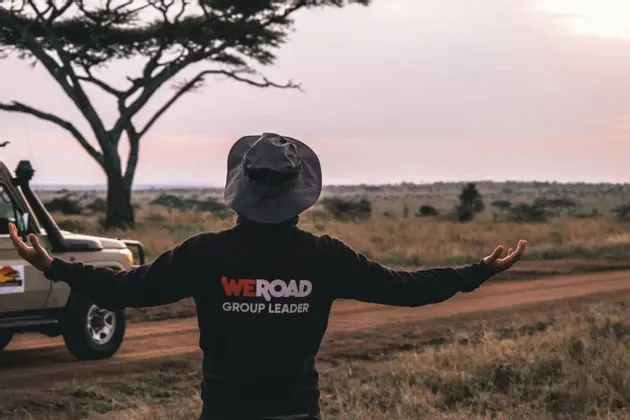 Ein WeRoad-Gruppenleiter steht, von hinten gesehen, mit ausgestreckten Armen in einer Savannenlandschaft neben einem Safari-Fahrzeug bei Sonnenuntergang.