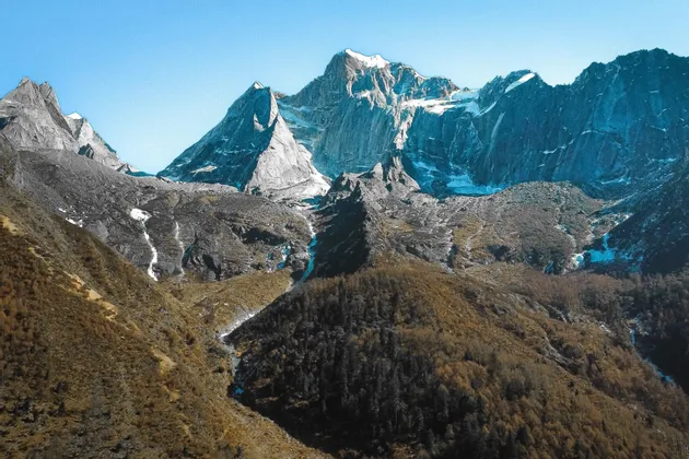Jagged, snow-capped mountains tower over a valley with autumn-colored forests under a clear blue sky.