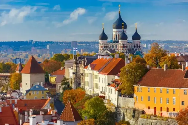 A cityscape featuring an Orthodox cathedral with black onion domes rising above colorful buildings and autumn trees under a blue sky.