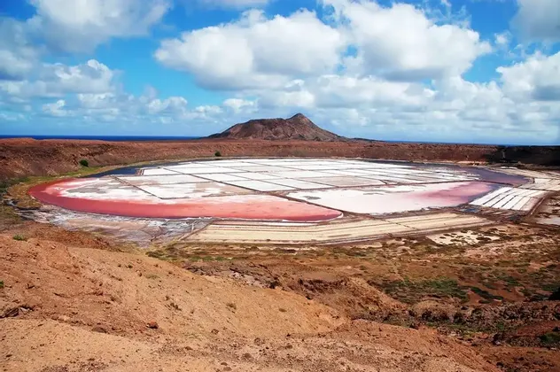 Saline rosa e bianche si estendono su un paesaggio arido e vulcanico, con una montagna e l'oceano visibili sullo sfondo.