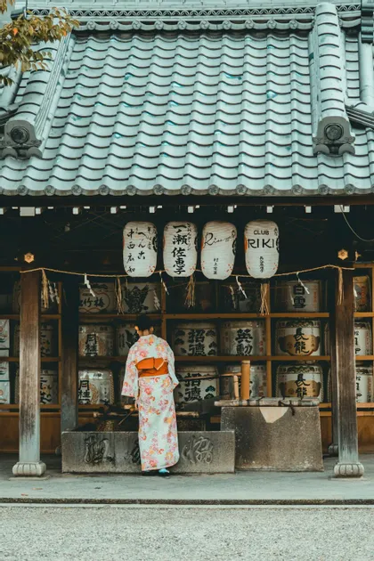 A woman in a floral kimono, seen from behind, stands at a water basin in front of a Japanese shrine with hanging lanterns.